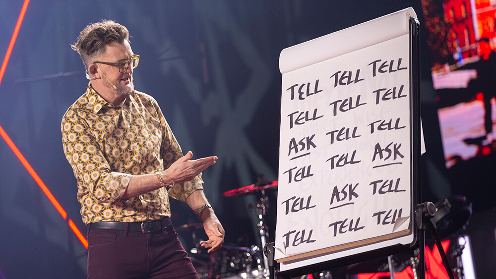 man speaking and pointing at sign with words 'ask' and 'tell' printed several times on it