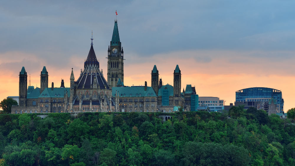 Canada’s Parliament buildings on a wooded hill above the Ottawa River at sunset, with the Peace Tower and the Canadian flag at centre.