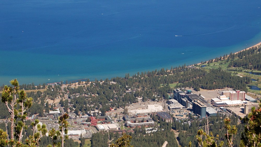 Aerial view of South Lake Tahoe from above, showing hotel towers along the shoreline and the lake stretching into the distance.